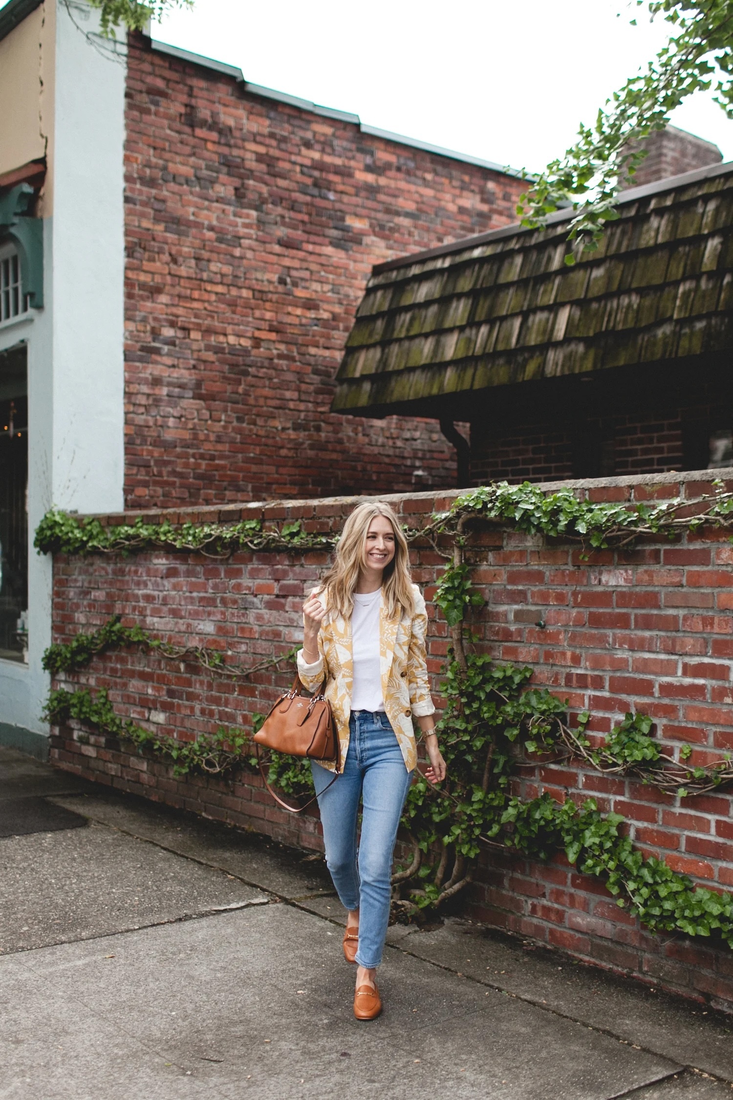 Person walking along a brick wall covered with green ivy, wearing a patterned jacket over a white top, blue jeans, tan shoes, and carrying a brown handbag
