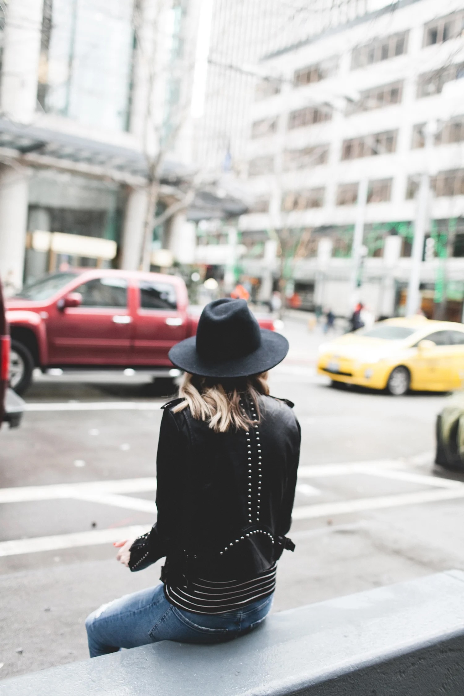 Person sitting on a city bench wearing a black wide-brim hat, black studded leather jacket, and striped top, with traffic and tall office buildings in the background.