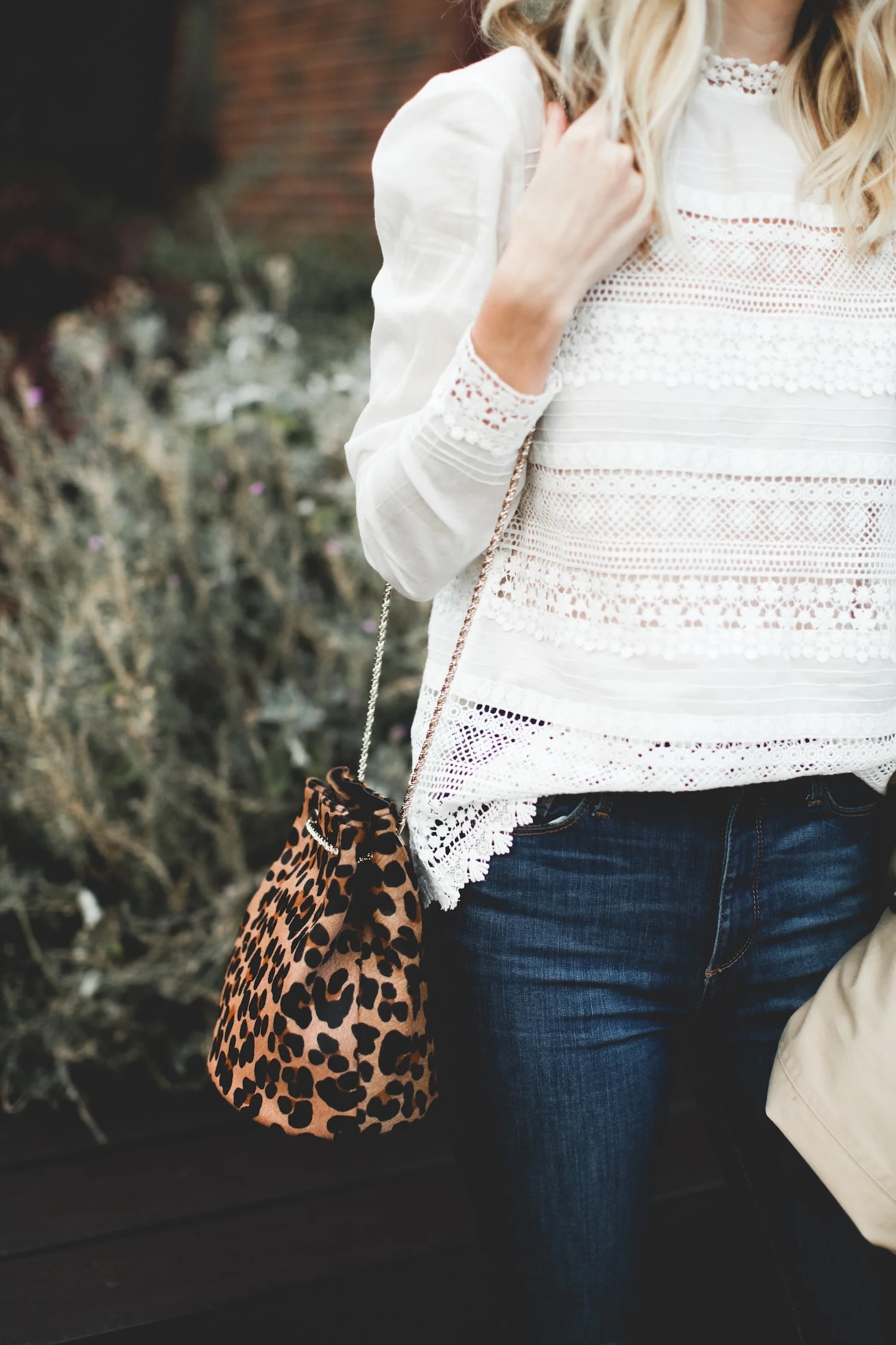 Close-up of a person wearing a white lace long-sleeve blouse paired with dark blue jeans, holding a small leopard-print bucket bag with a gold chain strap
